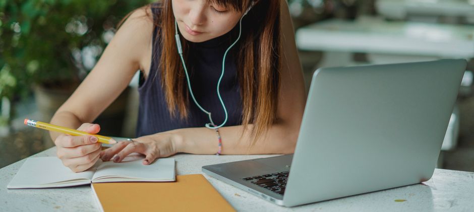person writing in notebook while listening to music with headphones and laptop on desk